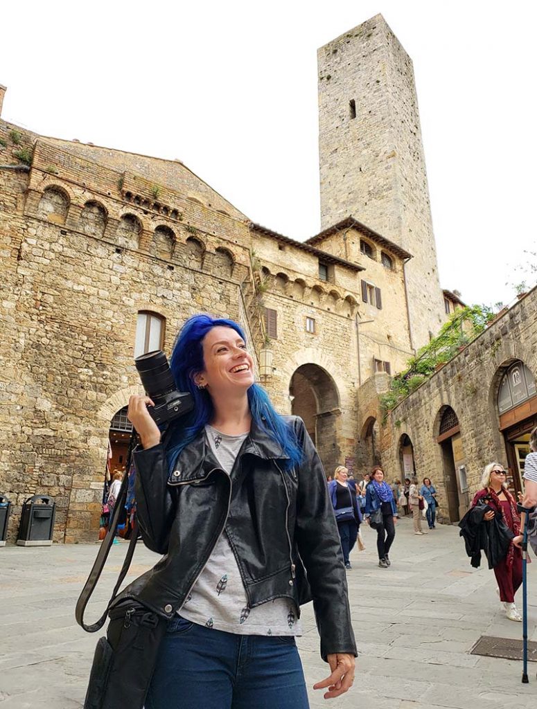 mulher segurando camera com uma mao ao lado de torre em san gimignano na italia