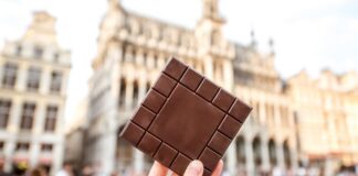 Roteiro do chocolate: 9 cidades europeias para os chocólatras de plantão Mão segurando barra de chocolate em frente a prédios históricos da Grand Place em Bruxelas, Bélgica